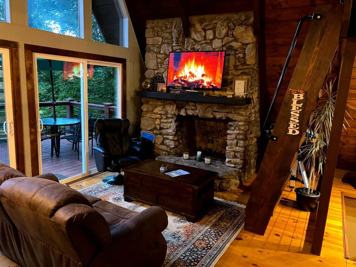 A close view of the main seating area with a plush sofa, coffee table, and crackling fireplace as the focal point. A television is mounted above the mantle, perfect for cozy movie nights or sharing stories after a day outdoors. The palette is warm and inviting, with tasteful decor and subtle nods to the A-Frame's character.