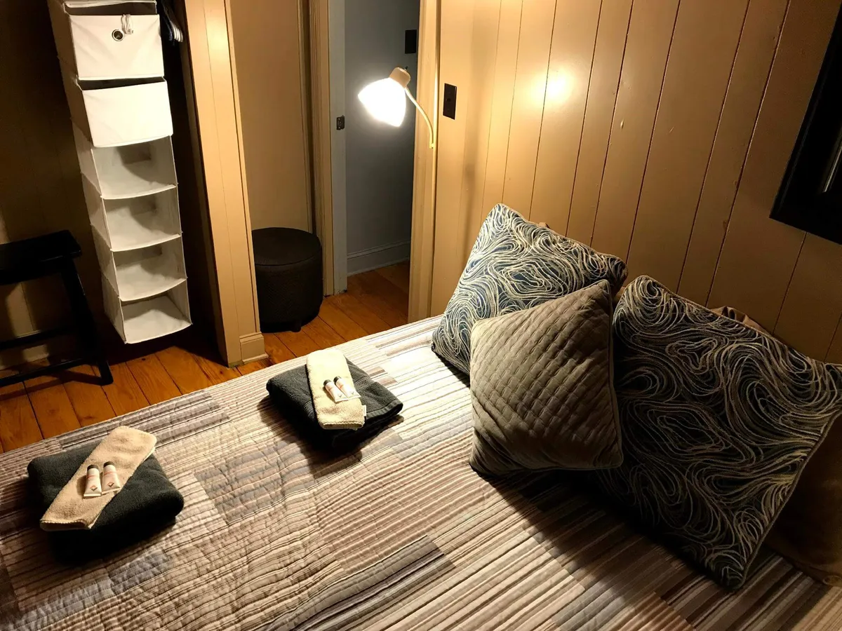 The room centers on a bed with plush pillows and neatly folded towels, paired with a floor lamp and a storage unit. The ambiance is intimate and softly lit, suggesting quiet evenings, restful sleep, and moments of relaxation.
