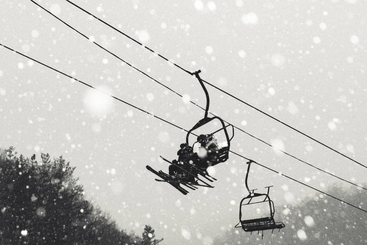 A scenic winter exterior featuring a ski lift gliding above a snow-covered landscape, with gentle snowfall creating a serene mountain atmosphere. The landscape evokes the excitement of skiing and snowboarding with the slopes clearly visible.