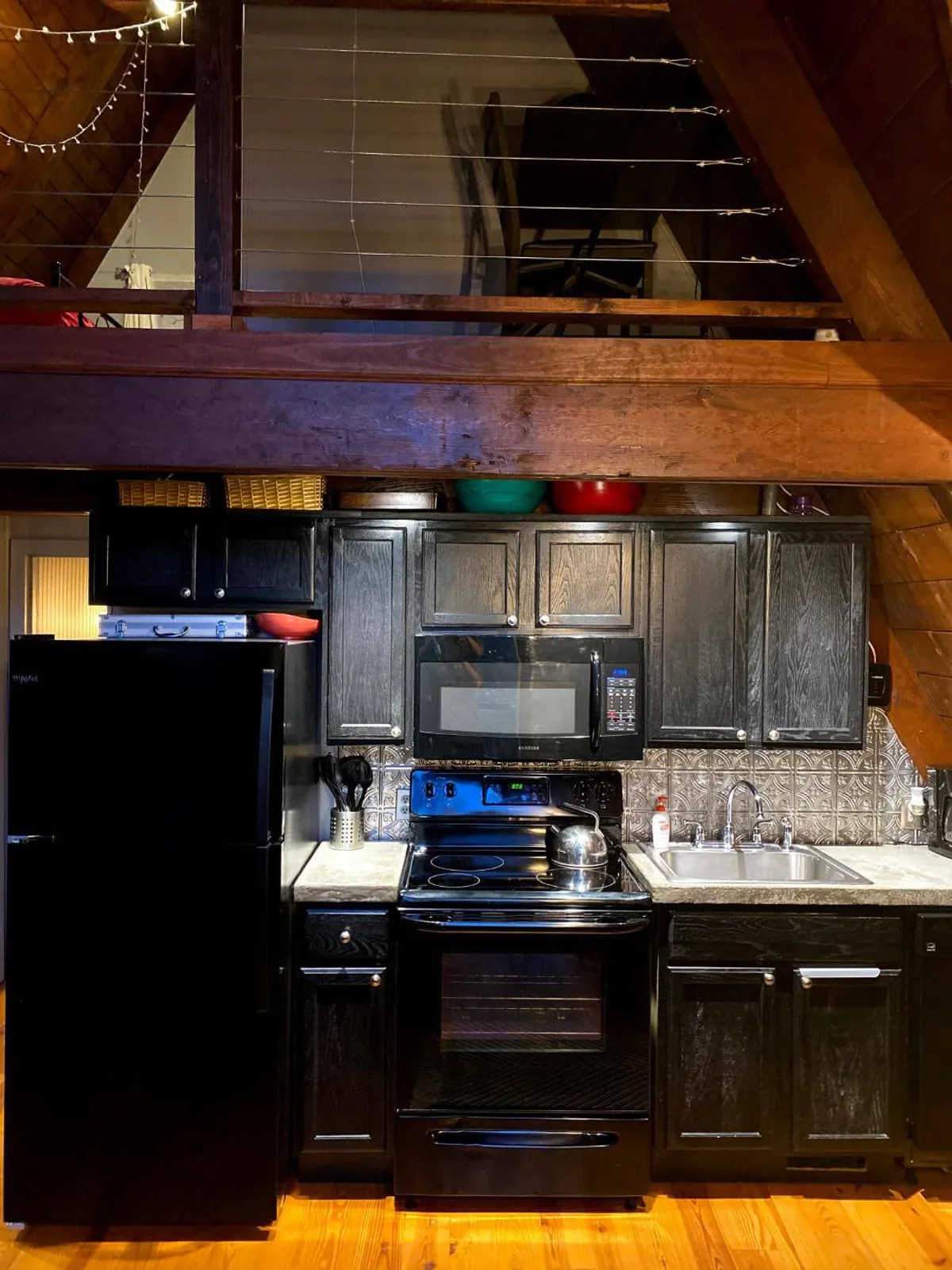 The photo showcases a cozy kitchen area with black appliances, wooden cabinetry, granite countertops, and decorative tile accents. The open loft space above hints at an expansive, sociable interior where cooking and gathering naturally blend together.