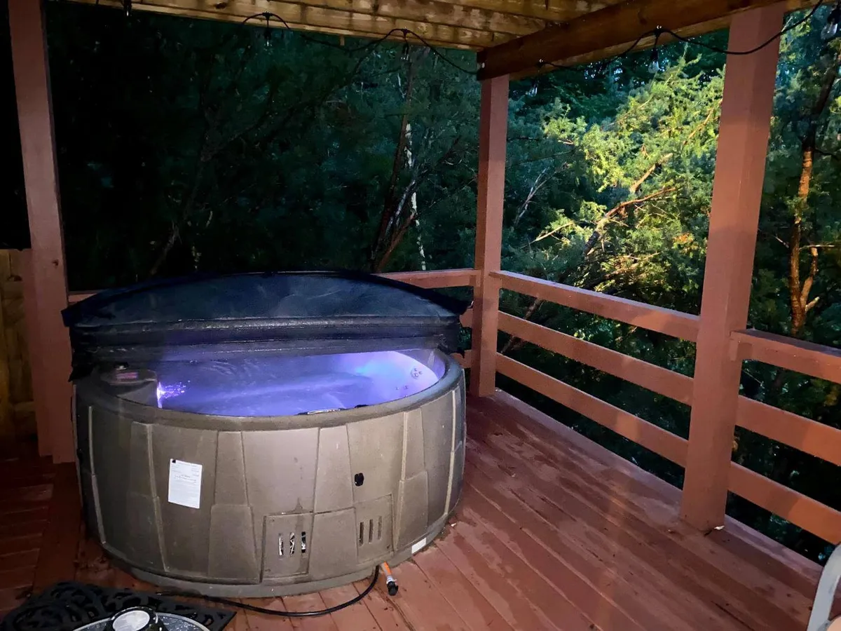 This image features an outdoor hot tub nestled within a wooden deck, surrounded by lush greenery and protected by a wooden railing. The atmosphere is serene, inviting guests to relax beneath the open sky and enjoy conversations or soak in the peaceful ambiance of nature.