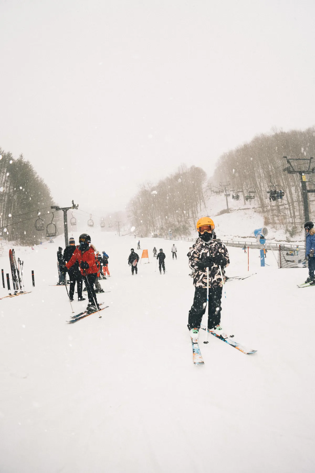 A lively winter scene with a skier in the foreground, a ski lift in the background, and a group of skiers and snowboarders on a snow-covered slope. The mood is vibrant, conveying the fun and camaraderie of outdoor snow sports for all ages.