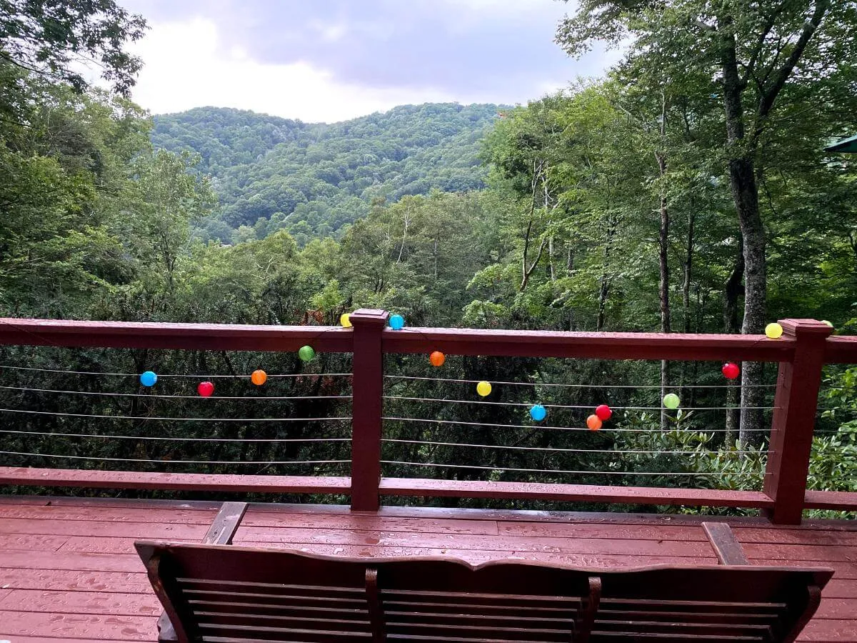 A view from a wooden deck adorned with colorful string lights, overlooking vibrant green mountain scenery. The scene suggests a tranquil spot for enjoying mountain breezes with a beverage, while the lights hint at cozy evenings and gatherings.