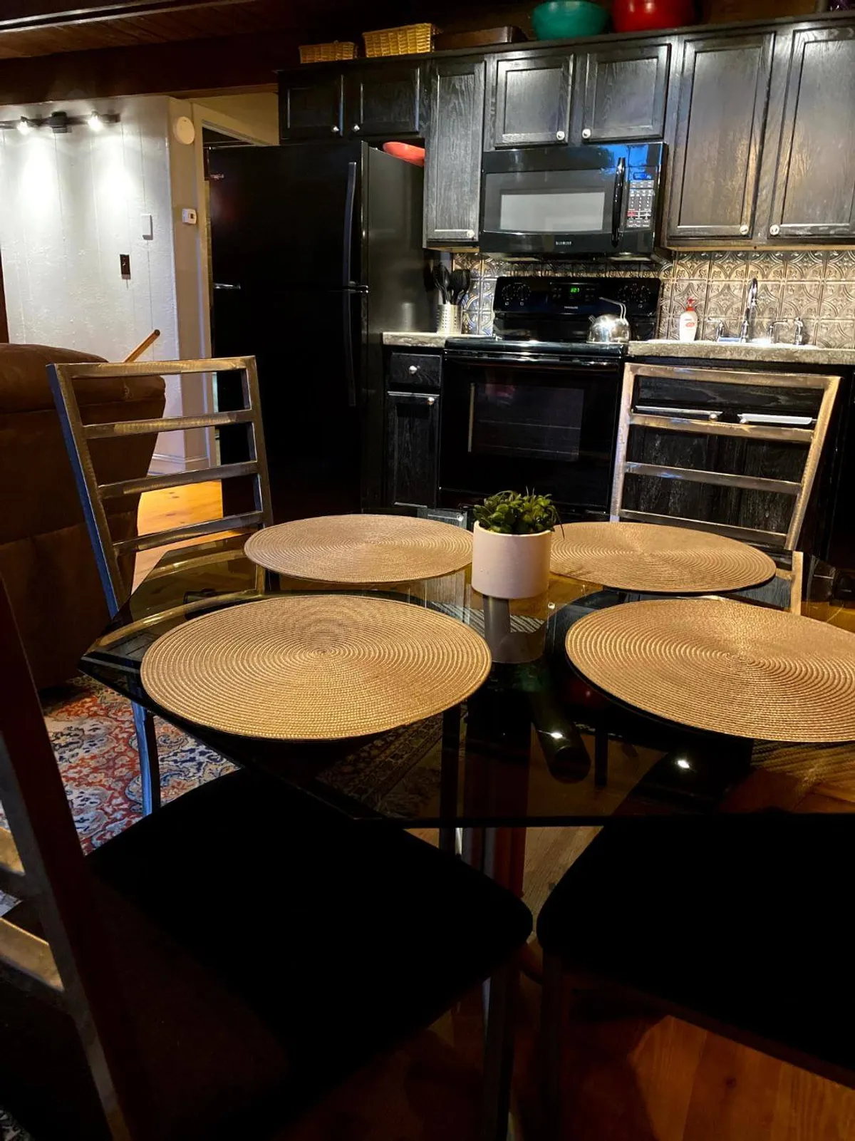 This image captures a stylish glass dining table set with woven placemats, a potted plant, and a view of kitchen appliances in the background. The dining area is warm and inviting, suited for both casual breakfasts and celebratory dinners.