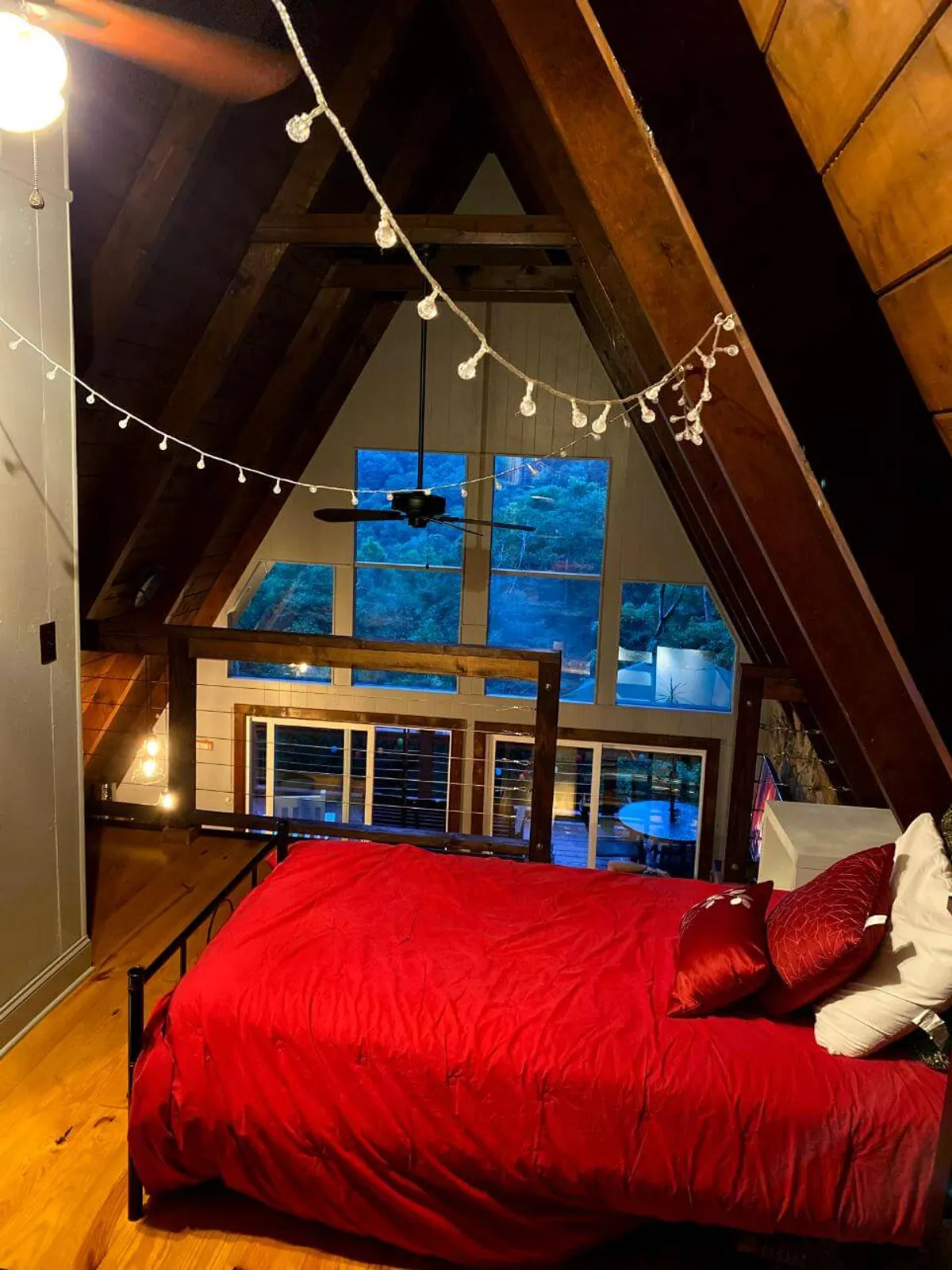 A comfortable bed with rich red bedding is set beneath wooden beams, flanked by string lights that add a touch of warmth and intimacy. Large windows overlook the surrounding outdoor area, inviting the natural world in and promising restful nights lulled by nature.