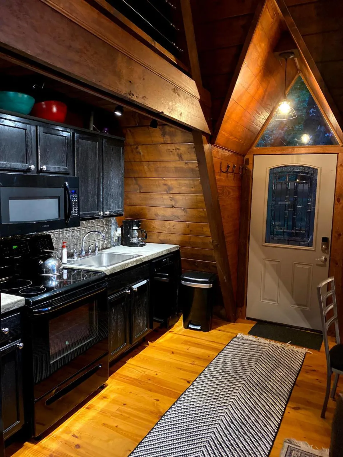 This image depicts a warmly lit kitchen with black appliances, exposed wooden beams, a kitchen sink, and a front door. The inviting setup suggests an ideal space for meal preparation and social interaction, capturing the heart of a communal cooking experience.