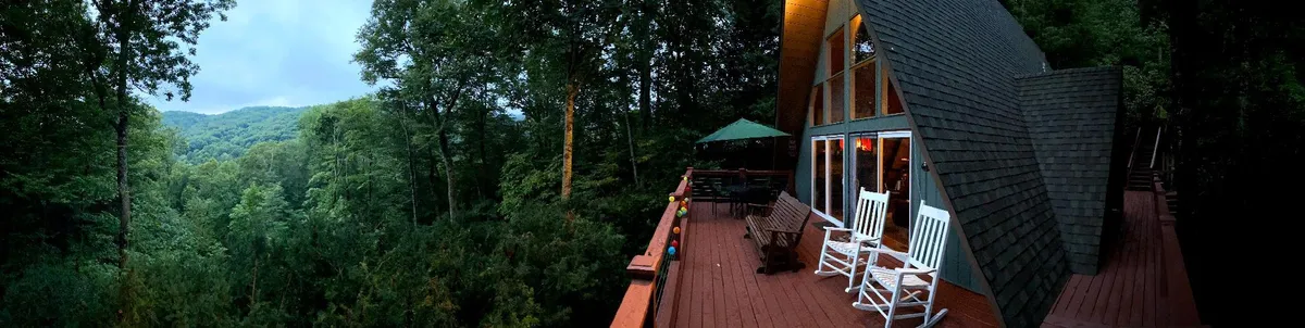 The hero image showcases the exterior deck of a distinctive A-frame cabin. Rocking chairs are arranged invitingly on a wooden deck, shaded by a green umbrella. The deck overlooks sweeping mountain views, with lush greenery stretching into the distance, evoking a sense of peace and seclusion. The scene is tranquil, perfect for relaxing, sipping coffee, or watching sunsets.
